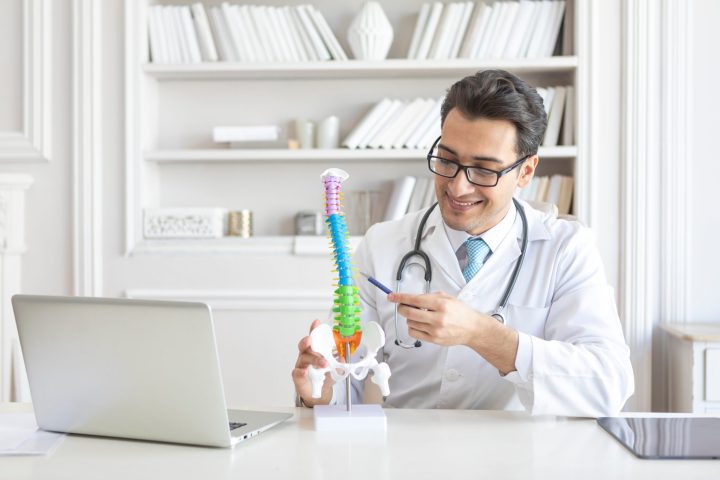 Young handsome male doctor orthopedist demonstrating the problem on spine bone model on the desk in his workplace